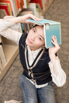 Playful young girl holding a blue book over her head near a bookshelf in library