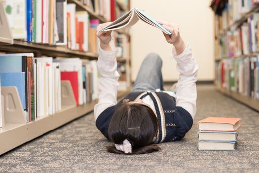 Young girl student laying on the library floor and reading a stack of books