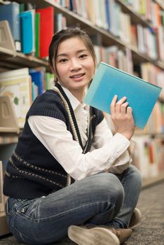 Young attractive woman holding a blue book while sitting in front of a bookshelf