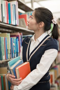 Attractive young female student holding a stack of books next to a bookshelf searching for books