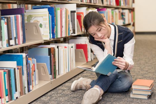 Young attractive woman reading a book while sitting on the floor in front of a bookshelf