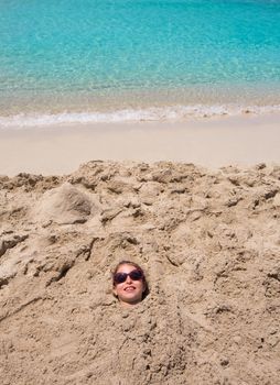 Funny kid girl playing buried in beach sand smiling with sunglasses at summer vacation