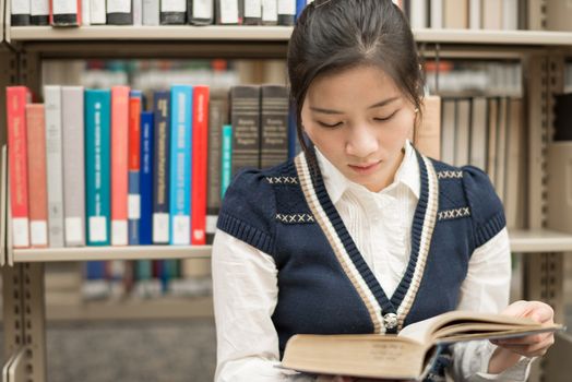 Attractive young girl sitting on the floor in front of a bookshelf reading a thick old book
