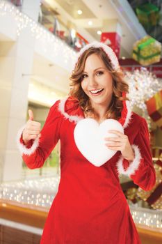 happy smiling Santa woman indoors, holding white heart, thumb up, background digitaly added, workpath