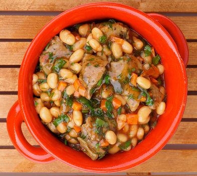 Delicious Homemade White Beans Stew with Beef and Greens in Red Pot closeup on Wooden background. Top View