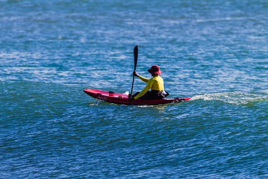 A man in a red hat rowing a red canoe in the ocean with a yellow life jacket on.