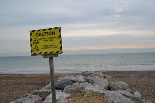 A yellow information sign warning of rocks and hidden objects along the seashore.Image taken along Worthing beach,Sussex,England.