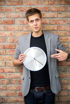 Young businessman holding a clock on brick wall background. Time in business concept