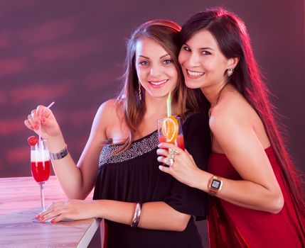 Two beautiful long haired female friends partying at a bar counter with cocktails in their hands under colorful lighting in a nightclub