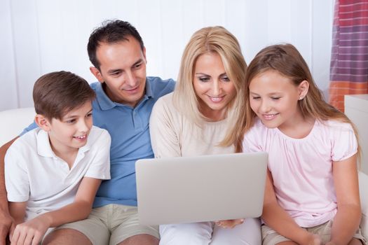 A Happy Family With Two Children Sitting On A Sofa Using Laptop At Home