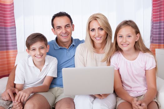 A Happy Family With Two Children Sitting On A Sofa Using Laptop At Home