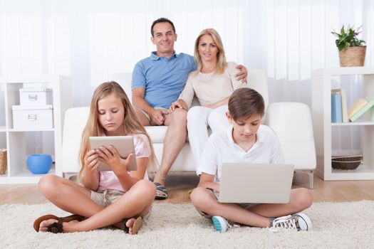 Children On The Carpet Using Tablet And Laptop With Parents Behind Them At Home