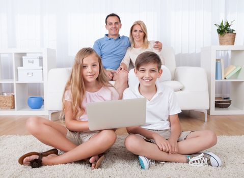 Children On The Carpet Using Tablet And Laptop With Parents Behind Them At Home