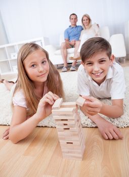 Children Playing With Wooden Blocks And Parents Behind Them At Home