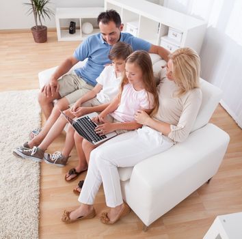 A Happy Family With Two Children Sitting On A Sofa Using Laptop At Home