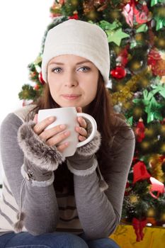 Woman in fur hat and mittens with mug under Christmas tree