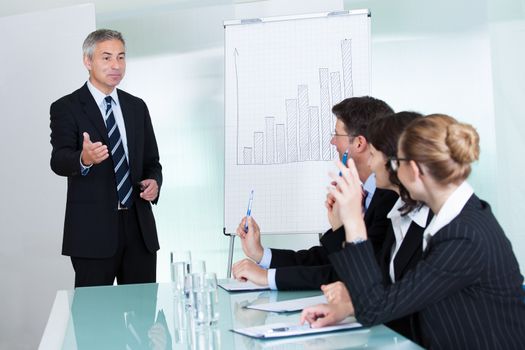 Manager or senior business executive standing in front of a graph giving a presentation to staff or colleagues seated at a table