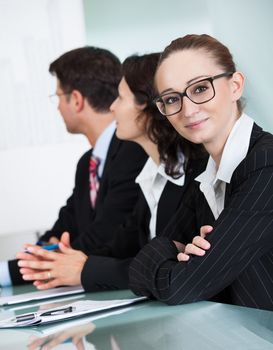 Beautiful young businesswoman with a charming smile wearing glasses looking at the camera while in a meeting with colleagues