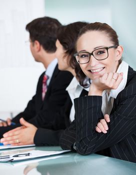 Beautiful young businesswoman with a charming smile wearing glasses looking at the camera while in a meeting with colleagues