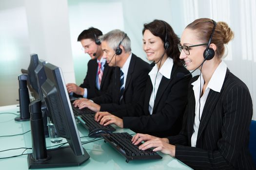 Line of professional stylish call centre operators wearing headsets seated behind their computers giving assistance