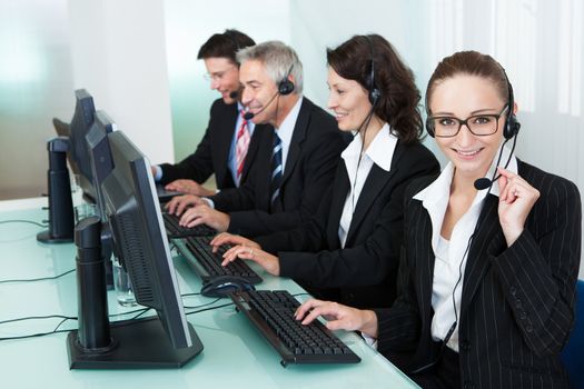 Line of professional stylish call centre operators wearing headsets seated behind their computers giving assistance