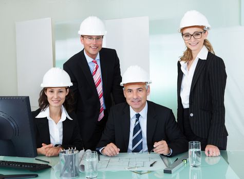 Four diverse professional partners in an architectural firm posing behind a table in an office wearing their hardhats and suits
