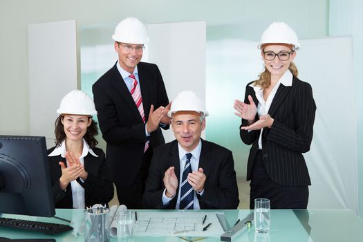 Team of four diverse architects or structural engineers wearing hardhats in an office applauding