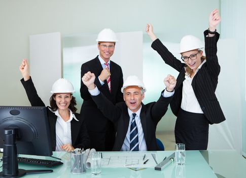 Team of four diverse architects or structural engineers wearing hardhats in an office cheering and raising their hands in the air