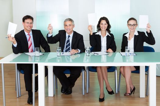 Group of four stylish professional judges seated at a long table holding up blank cards for their scores