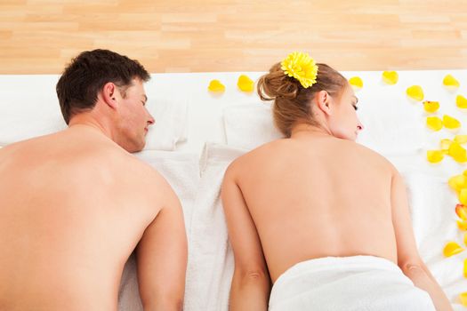 Smiling relaxed couple lying side by side on a table amongst yellow flowers at a spa with their top half bare and a towel around their loins