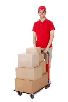 Cheerful young deliveryman in a red uniform holding trolley loaded with cardboard boxes isolated on white