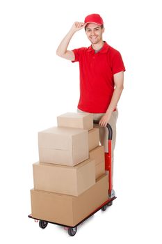 Cheerful young deliveryman in a red uniform holding trolley loaded with cardboard boxes isolated on white