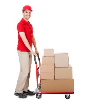 Cheerful young deliveryman in a red uniform holding trolley loaded with cardboard boxes isolated on white