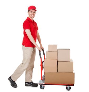 Cheerful young deliveryman in a red uniform holding trolley loaded with cardboard boxes isolated on white