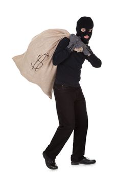 Thief in black clothes wearing a balaclava carrying a large bag of money with a dollar sign over his shoulder isolated on white