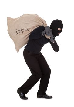 Thief in black clothes wearing a balaclava carrying a large bag of money with a dollar sign over his shoulder isolated on white