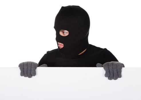 Thief in a balaclava and gloves looking around the edge of a blank sign with copyspace for your text isolated on white