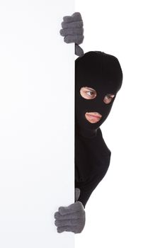 Thief in a balaclava and gloves looking around the edge of a blank sign with copyspace for your text isolated on white