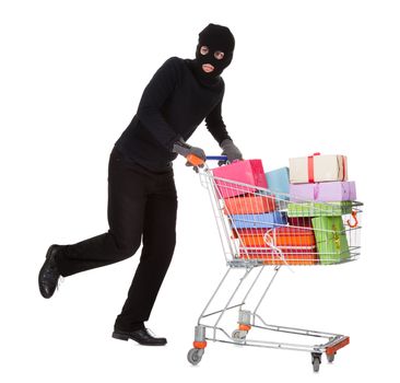 Thief in a black costume and balaclava pushing a shopping trolley full of gifts and presents from a celebration isolated on white