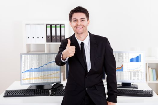 Confident smiling stock broker sitting on the edge of his desk surrounded by graphs and analytics indicating a successful bull market