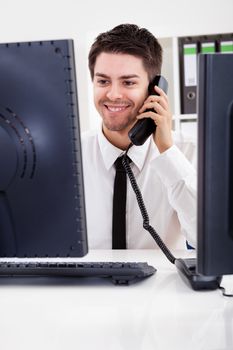 View between two computer monitors of a smiling handsome young stock broker talking on a phone