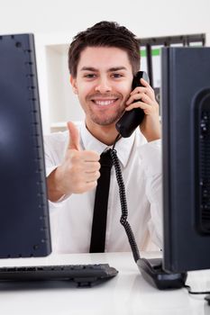 View between two computer monitors of a smiling handsome young stock broker talking on a phone