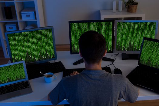 Rear high angle view of a male hacker sitting at a desk surrounded by computer monitors streaming data as he steals information