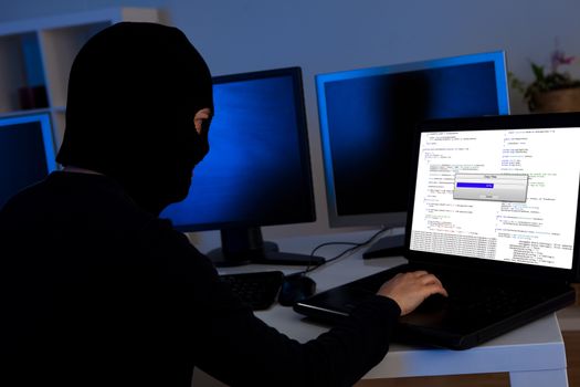 Masked hacker wearing a balaclava sitting at a desk downloading private information off a computer