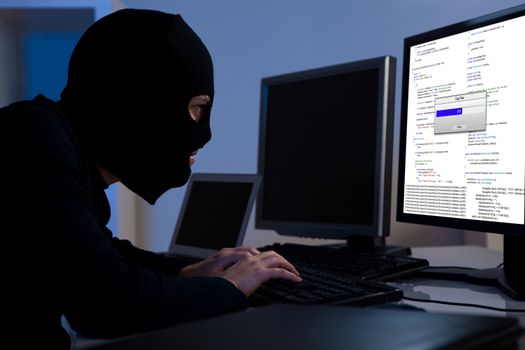 Masked hacker wearing a balaclava sitting at a desk downloading private information off a computer