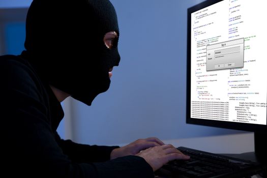 Masked hacker wearing a balaclava sitting at a desk downloading private information off a computer