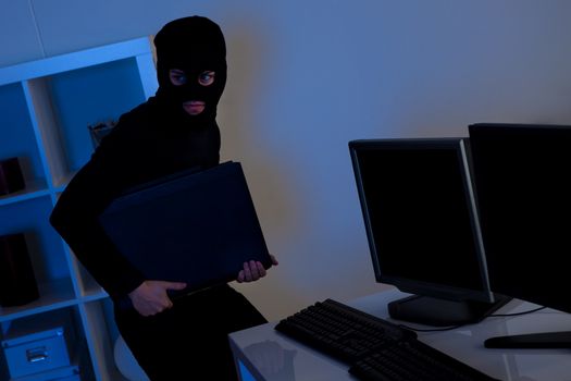Man dressed in black and wearing a balaclava is stealing a laptop from an office