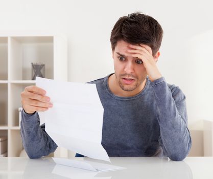 Horrified young man reading a document with an aghast expression and his hand to his forehead as he stares wide eyed at the page of paper