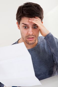 Horrified young man reading a document with an aghast expression and his hand to his forehead as he stares wide eyed at the page of paper