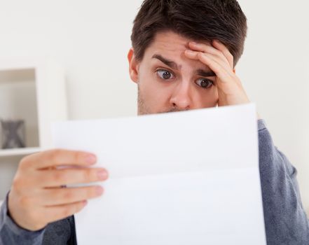 Horrified young man reading a document with an aghast expression and his hand to his forehead as he stares wide eyed at the page of paper